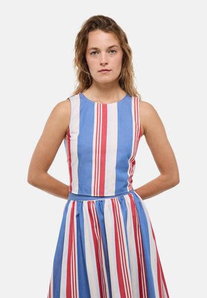 Jeune femme aux cheveux ondulés portant une robe sans manches à rayures verticales rouges, blanches et bleues, debout les mains dans le dos.