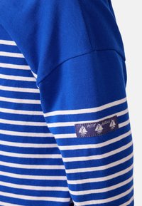 Langärmliges, blau-weiß gestreiftes Hemd mit rundem Ausschnitt, das ein Etikett mit kleinen Segelbootmotiven am Ärmel trägt.