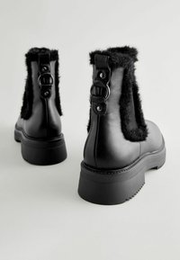 Botas de cuero negro con paneles laterales de peletería sintética, suela de goma ancha y detalles metálicos en la parte posterior, con un diseño de punta redondeada.