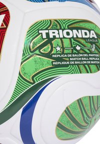 Biela futbalová lopta s akcentmi v zelené, modré a červenej farbe, na ktorej je nápis "TRIONDA League" a "Match Ball Replica" v niekoľkých jazykoch.