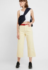 Crop topp i hvit ribbestrikket stoff, gule vide bukser med belte, og en marineblå genser drapert over skuldrene. Svartsneakers fullfører antrekket.