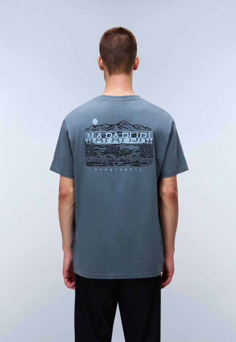 Jeune homme aux cheveux courts portant un T-shirt bleu-gris avec un paysage de montagne et le texte "Napapijri geographic" au dos.