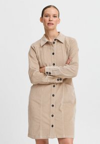 Beige kord/frotté klänning med krage, framknäppning med knappar, långa ärmar, sidofickor och subtil ribbad textur.