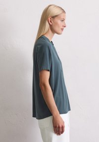 Türkise Bluse mit kurzen Ärmeln, lässiger Passform, Knopfdetails und weichem Material, kombiniert mit weißen Hosen.