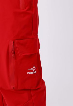 Gros plan sur la poche d'un pantalon cargo rouge avec fermeture éclair et rabat, arborant un logo PXG brodé en blanc sur le tissu.