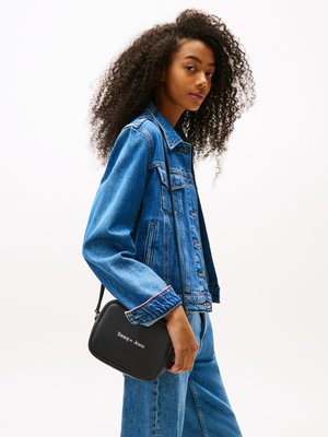 Sacoche noire à bandoulière de forme rectangulaire, avec une surface texturée et un logo brodé blanc. Portée avec une veste en denim bleue et un jean.