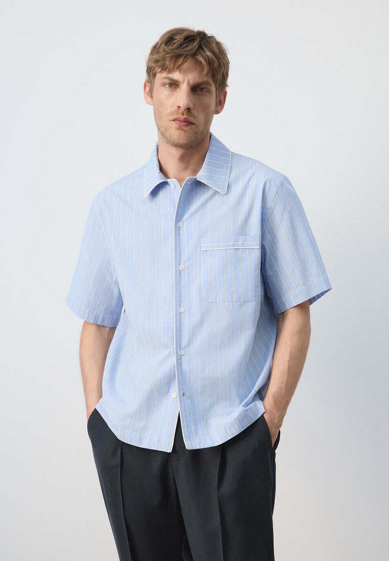 Homme aux cheveux courts et bruns portant une chemise bleu clair à manches courtes avec de fines rayures blanches et un pantalon foncé sur un fond neutre.