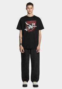 Czarna bawełniana koszulka z czerwono-białym graficznym wzorem tarczy i stylizowanym tekstem. Połączona z luźnymi czarnymi dżinsami i wielokolorowymi trampkami.