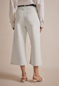 Weiße weite Jeanshosen. Merkmale sind zwei Gesäßtaschen, eine mittelhohe Taille und ein roher Saum. Getragen mit Riemchen-Absätzen.