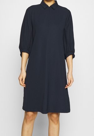 Mujer con un vestido azul marino suelto hasta la rodilla, con mangas abullonadas tres cuartos y cuello puntiagudo, sobre un fondo liso.