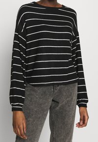 Schwarz-weißer, gestreifter Langarmoberteil mit weitem Ausschnitt und überschnittenen Schultern, kombiniert mit grau gewaschenen Jeans. Weiche Textur ist offensichtlich.