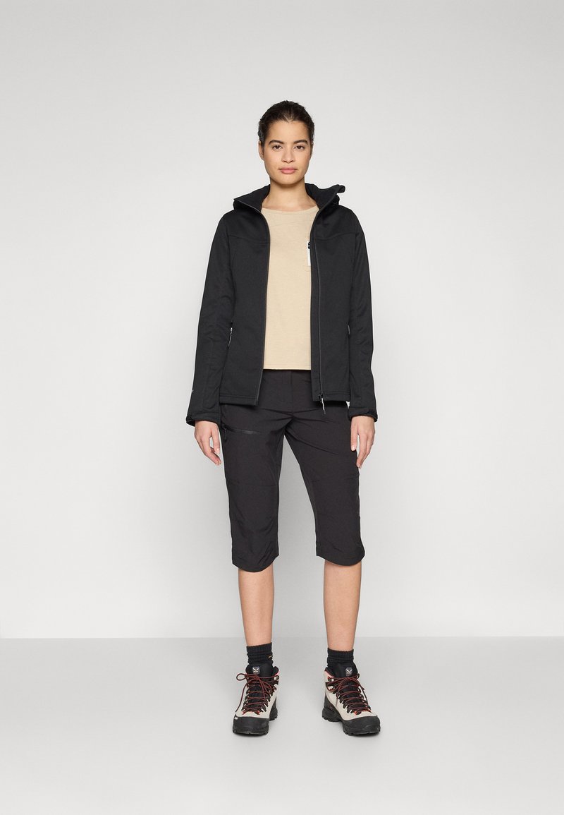 Chaqueta softshell negra con capucha, camiseta beige y pantalones cortos negros hasta la rodilla. Completado con botas de senderismo resistentes y calcetines oscuros.