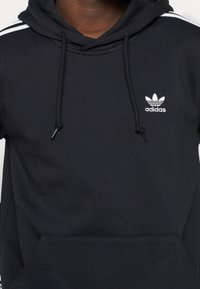 Musta puuvillainen huppari, jossa valkoinen Adidas-logo rinnassa ja valkoiset raidat olkapäillä. Edessä tasku.