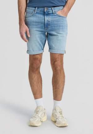 Mees kannab helesiniseid ülesrullitud teksashortse, valgeid kummisokke ja beeže tenniseid, seisab ühevärvilise tausta ees.