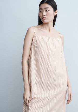 Νεαρή γυναίκα με μακριά σκούρα μαλλιά, που φοράει μπεζ και άσπρο ριγέ φόρεμα χωρίς μανίκια, κοιτάζει προς τα δεξιά της με φόντο ανοιχτό γκρι.