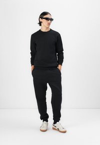 Completo felpa nera e pantaloni sportivi. Tessuto liscio, scollatura a girocollo, polsini a costine. Accompagnato da occhiali da sole neri e sneakers bianche da corsa.