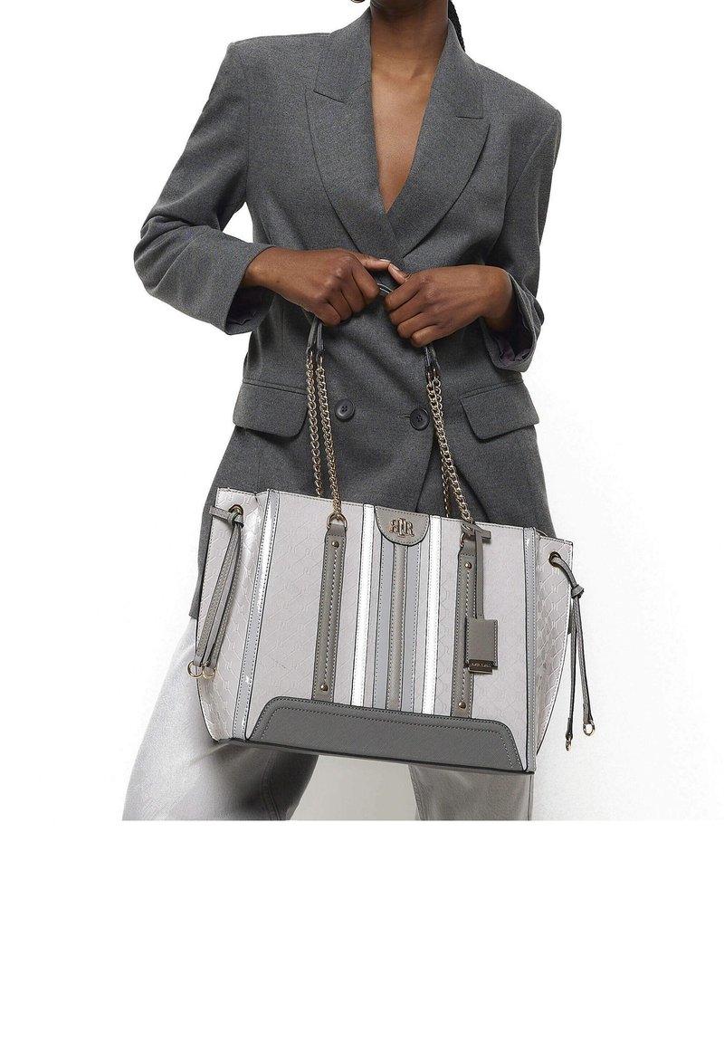 Personne portant un blazer gris tient un grand sac à main rayé gris et blanc avec des poignées en chaîne et des détails en cuir.