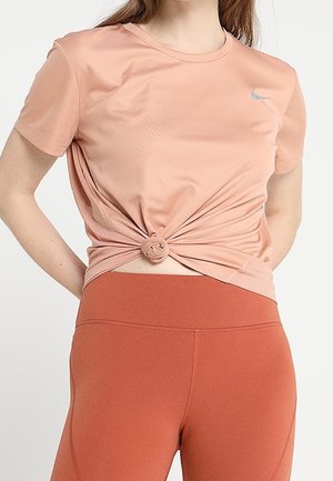 Kobieta nosząca jasnoróżowy krótki rękaw koszulki Nike, związany w talii, oraz wysokie legginsy w kolorze rdzy.