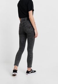 Pantalones vaqueros negros de talle alto con efecto desgastado, ajuste ajustado, diseño de cinco bolsillos y bajos deshilachados, combinados con zapatillas negras.