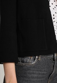 Cardigan noir en tricot avec une poche plaquée à l'avant, des manches trois-quarts, porté par-dessus un chemisier blanc à pois et associé à un jean en denim foncé.