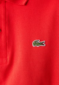 Camisa polo roja con una tela texturizada, que presenta un logotipo de cocodrilo bordado en verde en el lado izquierdo. Cuello con tapeta de dos botones.