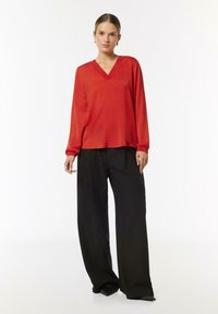 Rote Seidenbluse mit V-Ausschnitt und langen Ärmeln, kombiniert mit weiten schwarzen Hosen. Glatte Textur und minimalistisches Design.