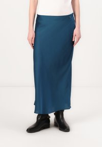Gonna maxi in satin teal con una texture liscia, caratterizzata da uno spacco laterale e un piccolo bottone argentato, abbinata a stivaletti neri.