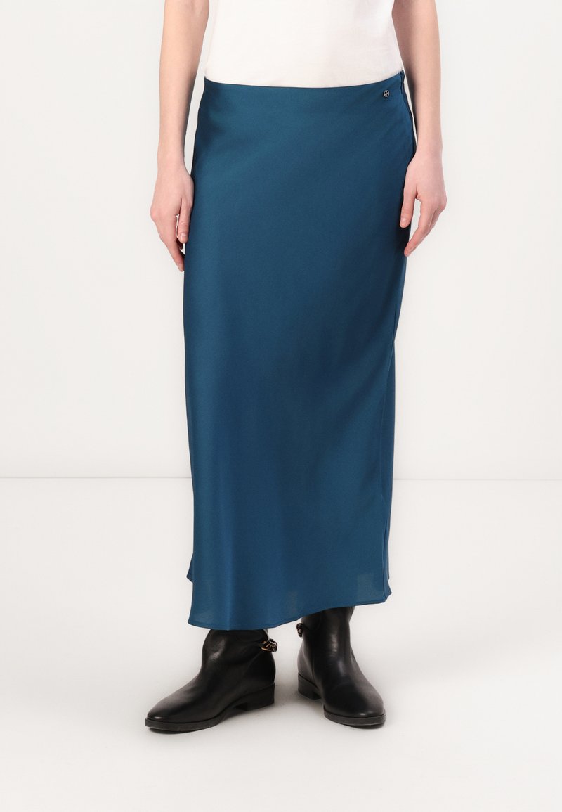 Gonna maxi in satin teal con una texture liscia, caratterizzata da uno spacco laterale e un piccolo bottone argentato, abbinata a stivaletti neri.