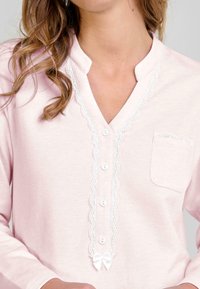 Mujer con una camisa de manga larga rosa claro, con encaje blanco en los bordes, botones en la parte delantera, un pequeño bolsillo con encaje y un lazo en la parte inferior.