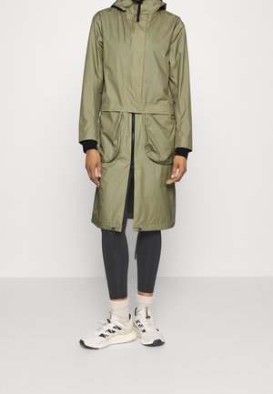 Persona con un impermeable largo con capucha de color verde oliva, leggings negros, calcetines blancos y zapatillas blancas, de pie frente a un fondo liso.