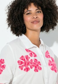 Weißes Hemd mit Knöpfen, das auf den Schultern mit leuchtend pinken Blumenapplikationen verziert ist, mit einer glatten Textur und entspannter Passform.