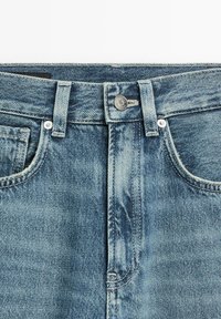 Lichtblauwe spijkerbroek met een tailleband met knopen, riemlussen, voorzakken en zichtbare stiksels op een witte achtergrond.