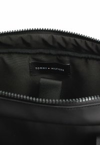 Interno di una borsa in nylon nera con una texture liscia, dotata di chiusura con zip e un'etichetta "TOMMY HILFIGER" ben visibile sulla fodera.