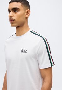 Νεαρός άνδρας που φοράει λευκό T-shirt EA7 Emporio Armani με μαύρη, πράσινη και κόκκινη ρίγα σε ταινία στους ώμους, σε απλό φόντο.