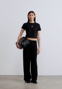 Μαύρο κοντομάνικο t-shirt με λογότυπο, σε συνδυασμό με μαύρο παντελόνι φαρδιάς γραμμής. Το μοντέλο κρατάει μια μαλακή μαύρη δερμάτινη τσάντα. Μινιμαλιστικός σχεδιασμός, λεία υφή.