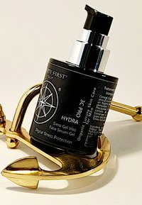 Zwarte fles met gezichtserum gel met een zilveren pompkop, geplaatst op een gouden anker. Het etiket bevat de productnaam en beschrijving in witte tekst.