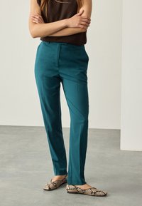 Pantalones verde azulado con textura suave, corte recto y pliegues delanteros, combinados con top marrón sin mangas y zapatos planos con estampado.