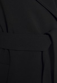 Schwarzes Stoffkleidungsstück mit glatter Textur, das eine taillierte Taille und einen Reverskragen aufweist. Die Nähte sind entlang der Kanten sichtbar.