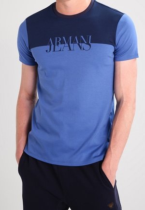 Mężczyzna w niebieskiej koszulce z krótkim rękawem i ciemnoniebieskim karczku, z logo "Armani Jeans" na piersi, z rękami w kieszeniach, na jednolitym tle.