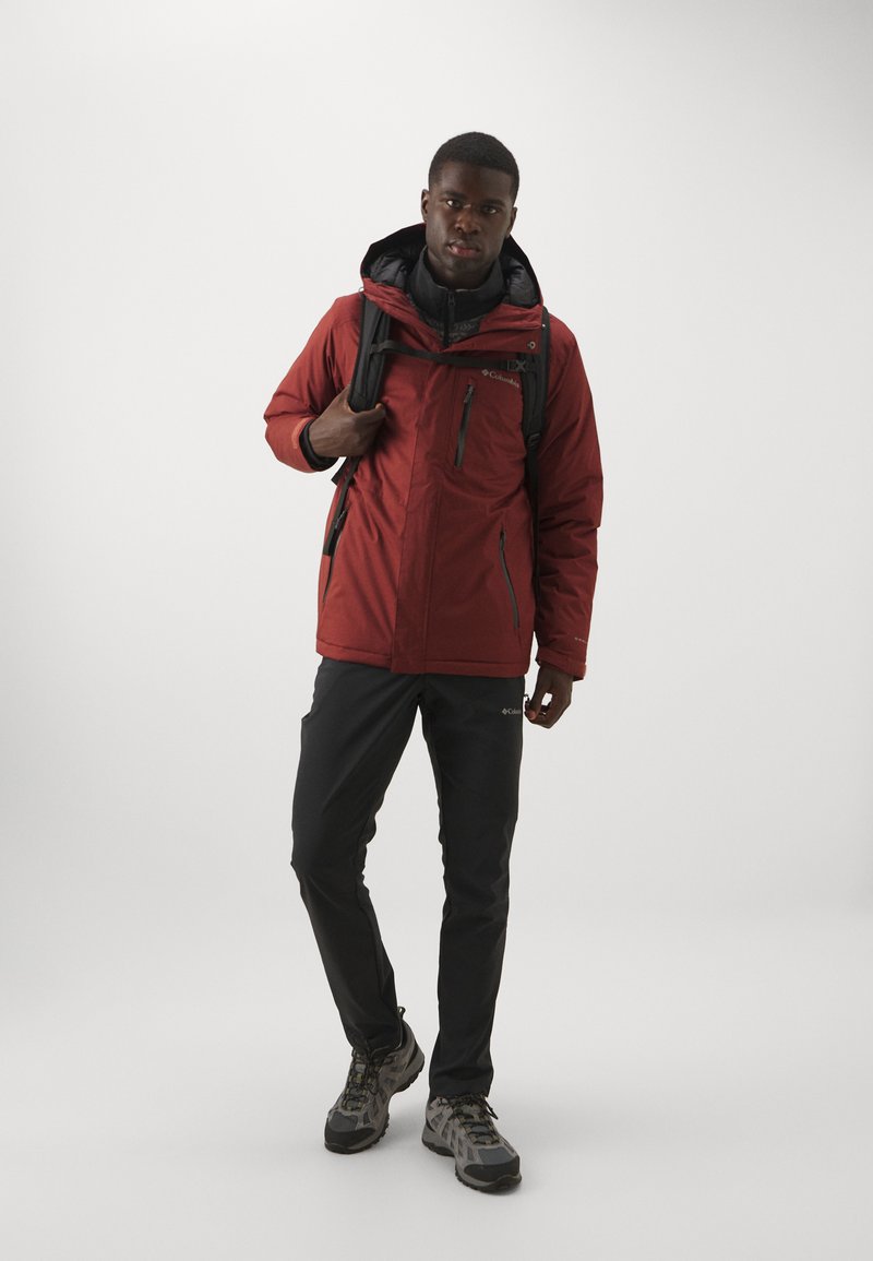 Chaqueta aislante roja con detalles negros, bolsillos con cremallera y capucha ajustable; combinada con pantalones negros y zapatos de senderismo grises.