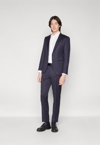 Costum bleumarin cu croială ajustată, jachetă cu un singur rând de nasturi, rever cu ureche și pantaloni fără pliuri. Asortat cu o cămașă albă și pantofi negri.
