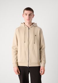 Felpa con zip beige dal finish testurizzato, cappuccio regolabile con cordino, chiusura frontale con zip e polsini a costine. Design minimalista con piccolo logo.
