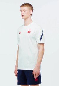 Camisa deportiva de manga corta blanca con logo de gallo rojo y rayas. Emparejada con pantalones cortos azul oscuro que presentan un logo a juego.