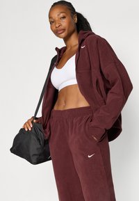 Bordo fleece jakna s patentnim zatvaračem, u paru s odgovarajućim trenirkama. Uključeni bijeli sportski grudnjak i crna torba za rame s Nike logotipom.