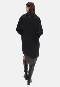 Zwarte oversized jas met een kraag, lange mouwen en een soepele textuur, gedragen over een donkergrijze jurk en knielange zwarte laarzen.