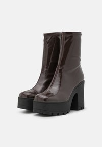 Bottes à plateforme marron foncé brillantes avec talons hauts épais et semelles noires épaisses et robustes sur fond blanc.