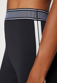 Melnas sporta legingi ar sānu baltām svītrām, ar teksturētu melnu un zilu svītrojumu jostas daļā, rādot tuvu attēlu uz jostas vietu.