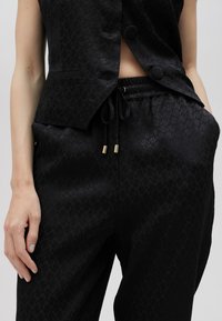 Svart matchande set som inkluderar en figurnära, ärmlös blazer med knappar och avslappnade byxor med dragsko och guldaccenter.