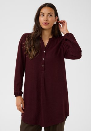 Mujer con el pelo largo y castaño lleva una túnica burdeos de manga larga y pantalones marrones, toca su pendiente y mira hacia un lado.