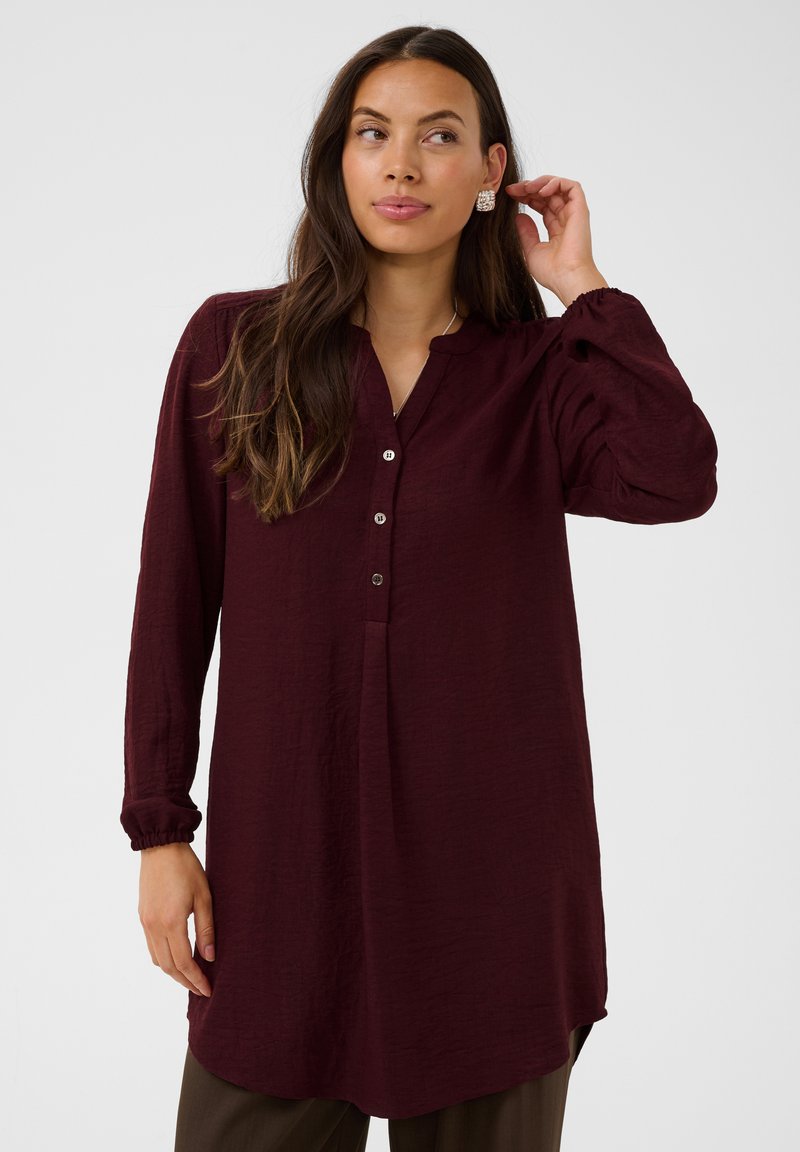 Femme aux longs cheveux bruns portant une tunique bordeaux à manches longues et un pantalon brun, touche sa boucle d’oreille et regarde sur le côté.